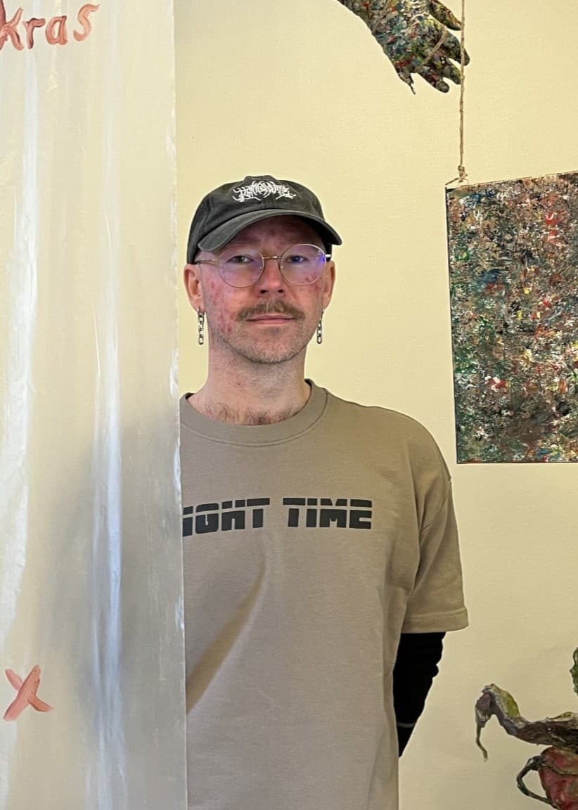En konstutställning med en man i beige t-shirt och keps stående bredvid abstrakta konstverk, inklusive en färgglad målning och en skulptur. En handskriven text syns på glasvägg i förgrunden.