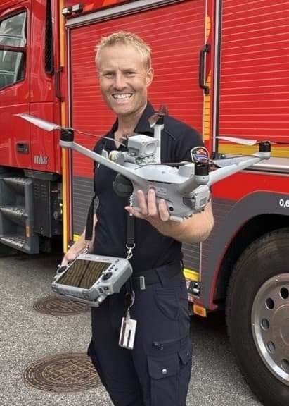 En brandman, identifierad som Daniel Eggertsen, står framför en röd brandbil och håller en drönare i handen. Drönaren används för räddningstjänstens insatser och övervakning.