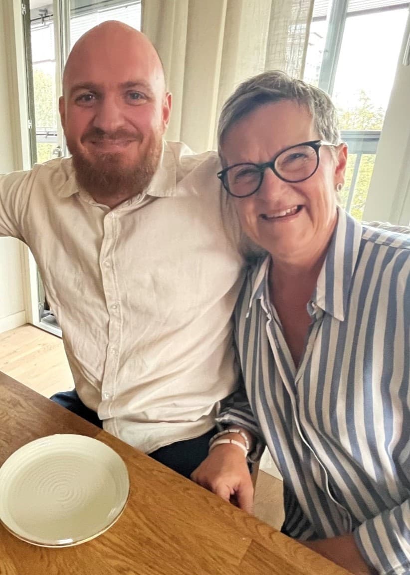 Tre personer, Per Thyberg, Alexander Wing och Inger Thyberg, sitter vid ett träbord med en kopp kaffe och en tom tallrik framför sig. De ser ut att vara i ett ljust kök med stora fönster och utsikt över grönska. Alexander kramar om Per och Inger och alla ler mot kameran. 