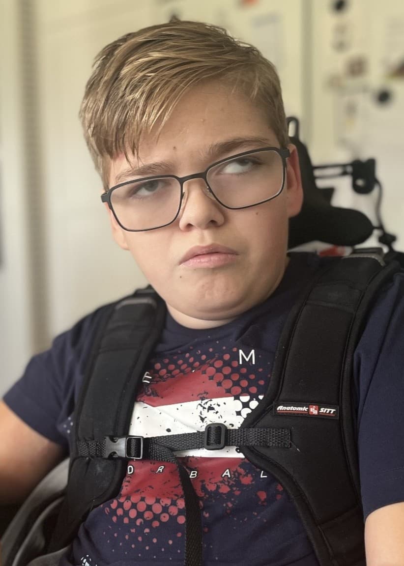 Elis Appelgren, 15, sitter i en rullstol vid ett köksbord. Han har en mörkblå t-shirt och omgivningen visar ett kök med vit inredning.