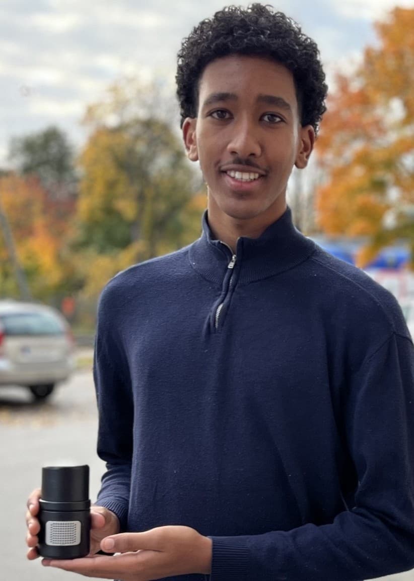 William Assefaw står utomhus på en höstig gata, klädd i blå tröja och jeans, och håller i en lampa. Bakgrunden visar parkerade bilar, träd med höstlöv och en byggnad i fjärran.