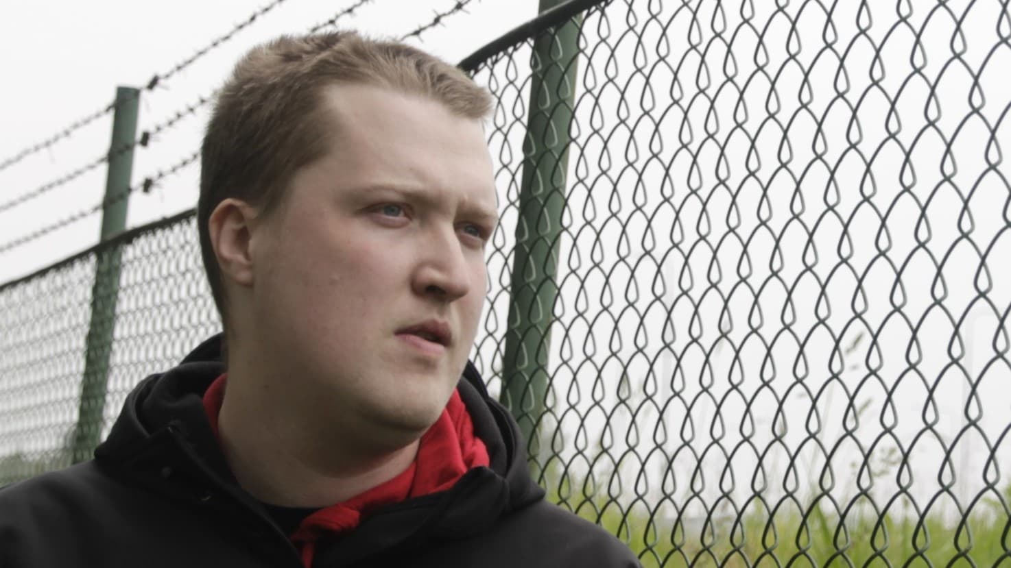 Eric Stellemark står vid ett stängsel i en utomhusmiljö med gräs och dimmig bakgrund. Han är klädd i en mörk jacka med en röd detalj.