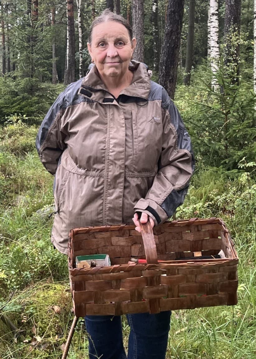 Evy Andreasson, svampkonsulent, står klädd i ljusbrun friluftsjacka i en grönskande skog med en korg fylld av svamp. 