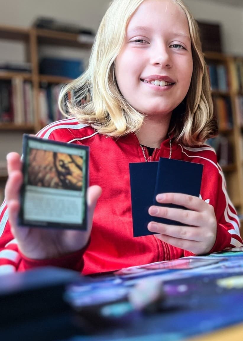 Blond flicka sitter vid ett bord i ett bibliotek och spelar ett kortspel med fantasy-tema. Hon håller upp ett spelkort i handen, och bordet är täckt av kort och spelmaterial.