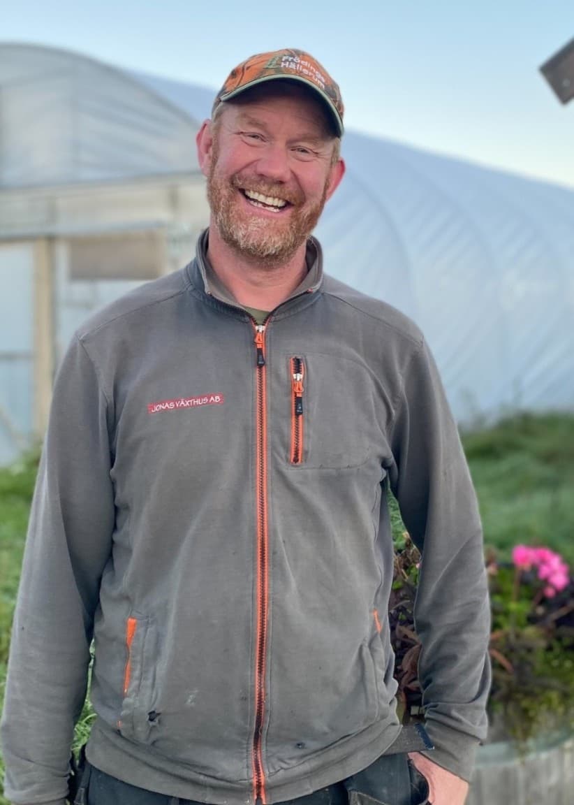 Jonas Enderberg står framför ett växthus på en grönskande gård, klädd i en grå jacka, med gräs och en skylt i trä intill sig.