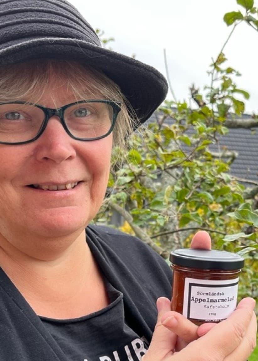 Karin Liljegren håller upp en burk med marmelad framför ett fruktträd i en trädgård. Hon bär en svart hatt och står omgiven av grönska.