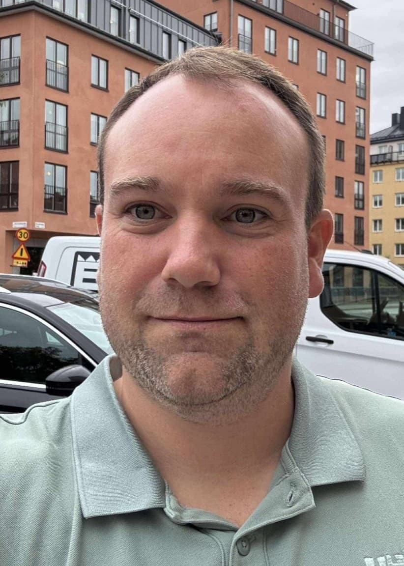 Mikael Nyrén framför en gata med parkerade bilar och flerbostadshus i bakgrunden. Molnigt väder och urban miljö med trottoar och vägskyltar synliga.