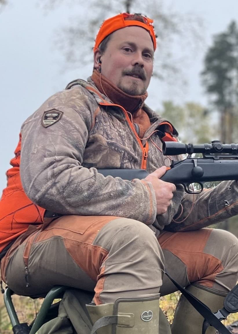 Jägaren Gustav Källsand sitter på en stubbe i skogen, klädd i jaktkläder och orange varselmössa, med ett gevär i händerna. Omkring honom syns höstfärgad natur och tätt skogsparti.