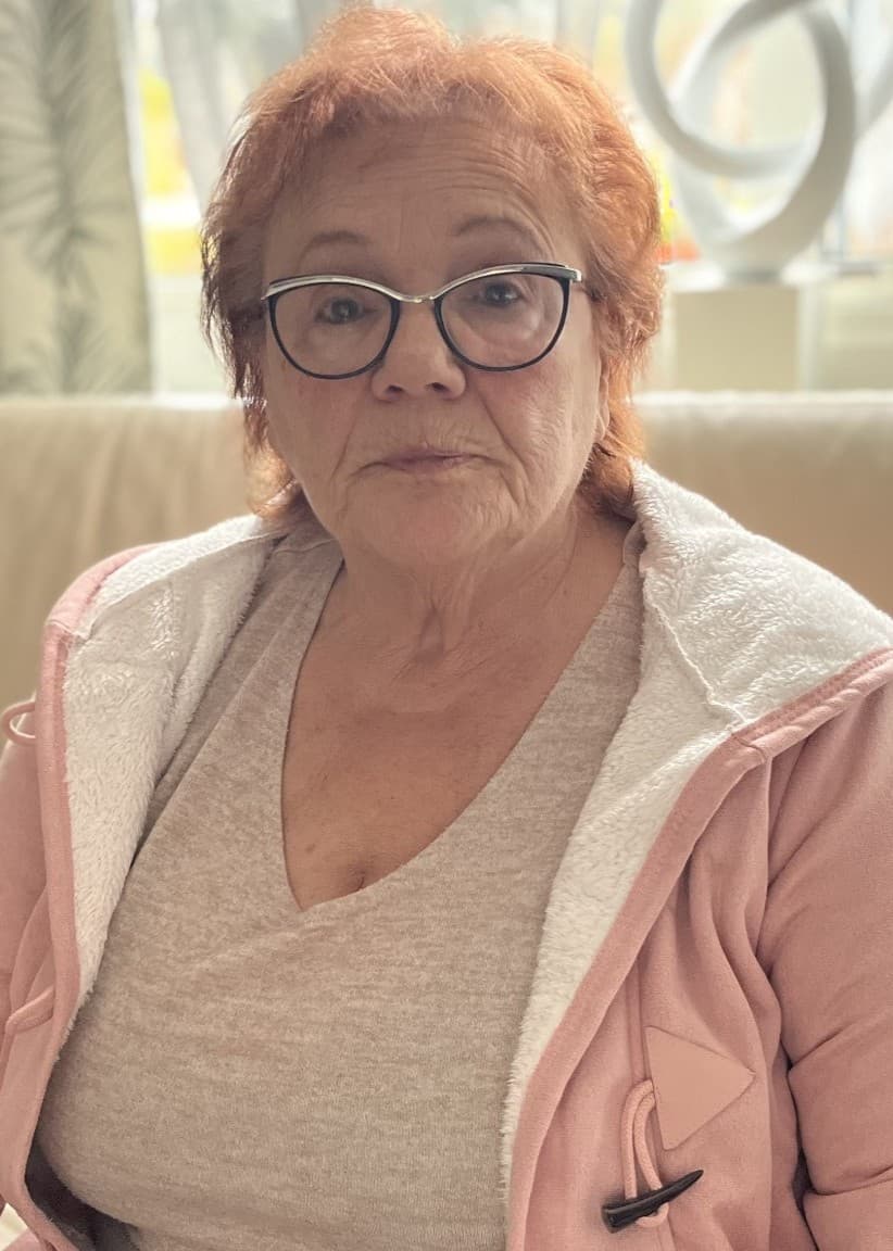 Zorica Grujic sitter på en ljus soffa med färgglada kuddar i bakgrunden. Hon håller i en rosa jacka och har ett bandage på ena armen. Rummet har en ljus inredning.