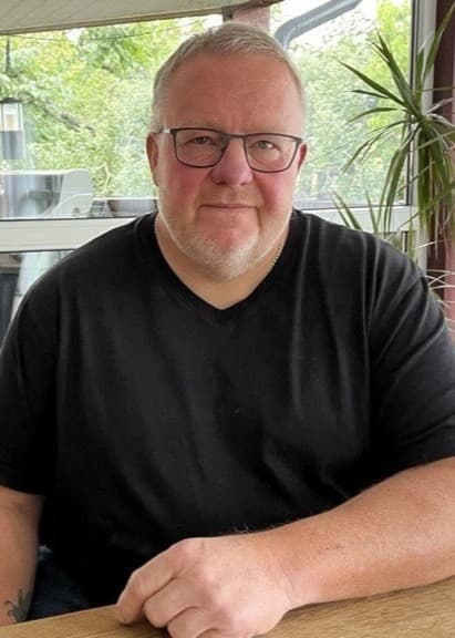 Jörgen Hall sitter vid ett bord på en terrass med glasväggar och grönska i bakgrunden. Han bär en svart t-shirt och omgivningen ger en lugn atmosfär.
