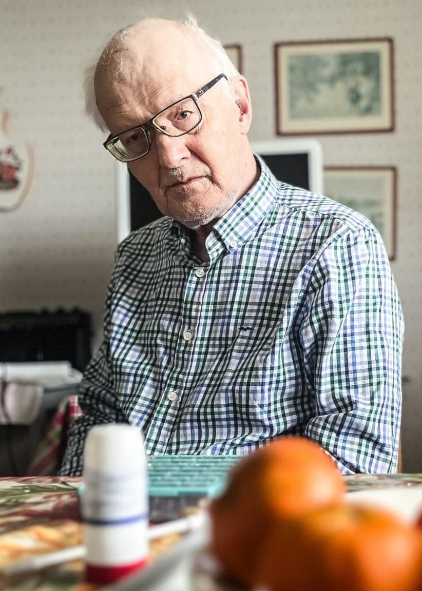 Göran Dalin sitter vid ett köksbord med mandariner och medicin i förgrunden. Bakgrunden visar en köksinteriör med väggdekorationer och en mikrovågsugn.