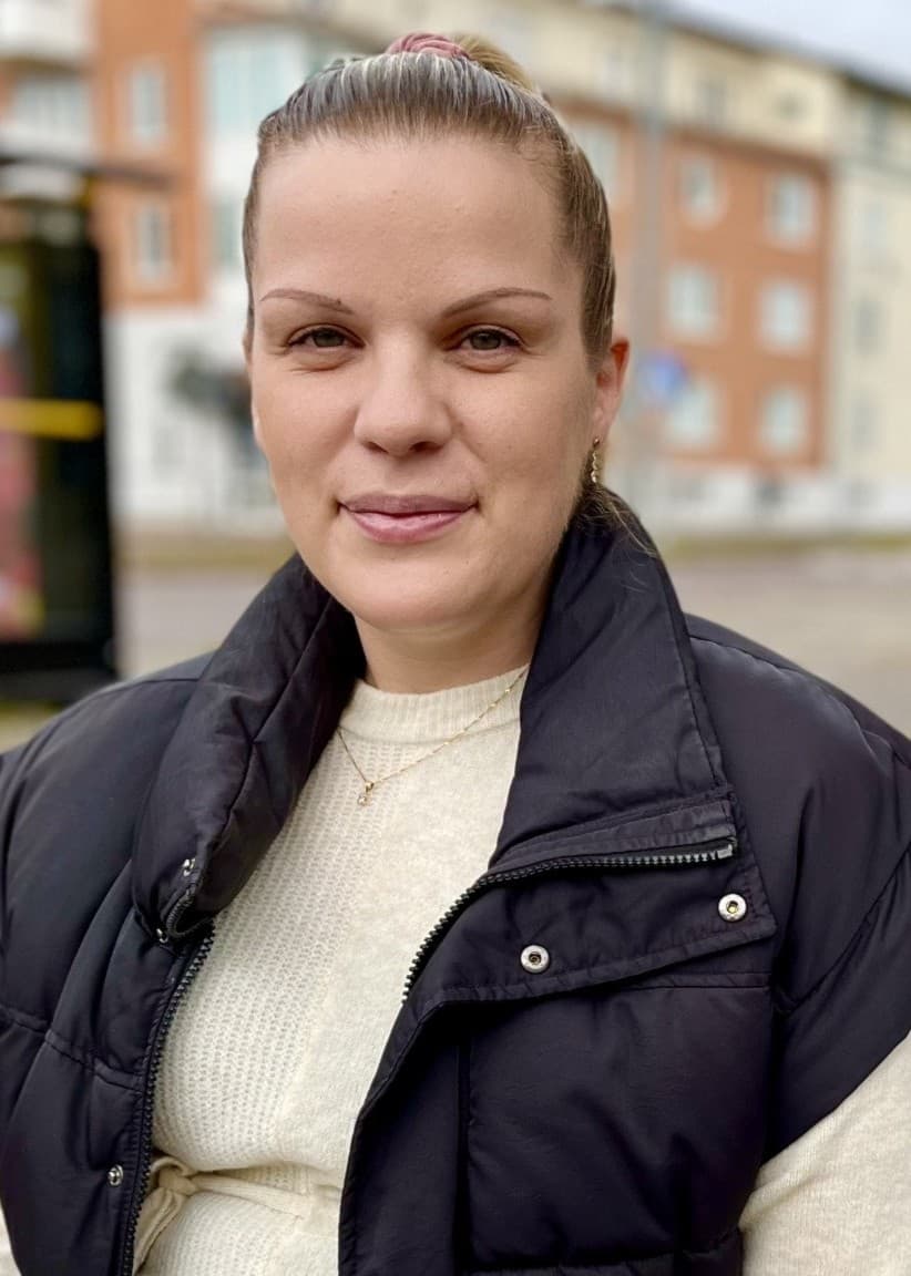 En kvinna sitter vid ett träd nära en busshållplats. Bakgrunden visar flerfamiljshus och en gångväg med grus och sten runt trädet.