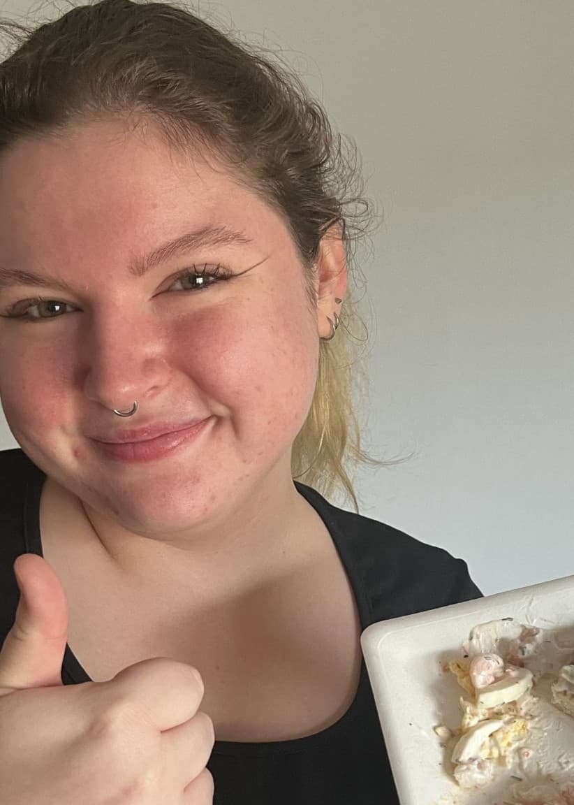Rebecca Dennisdotter håller upp en tallrik med mat, inklusive skinka, ost och grönsaker, och visar en tumme upp. Bakgrunden är enkel och neutral med en vit vägg.