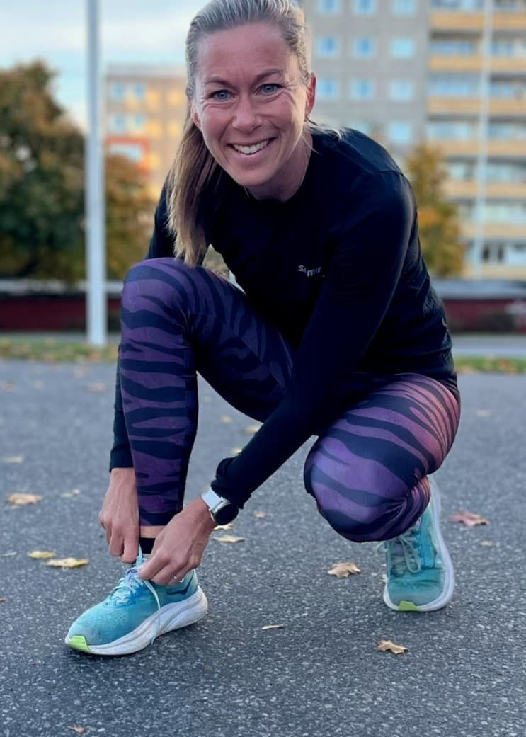 Frida Södermark, löpare och träningsinspiratör från Norrköping, ses knyta sina löparskor på en asfalterad yta. I bakgrunden syns höghus, höstfärgade träd och parkeringsplatser.