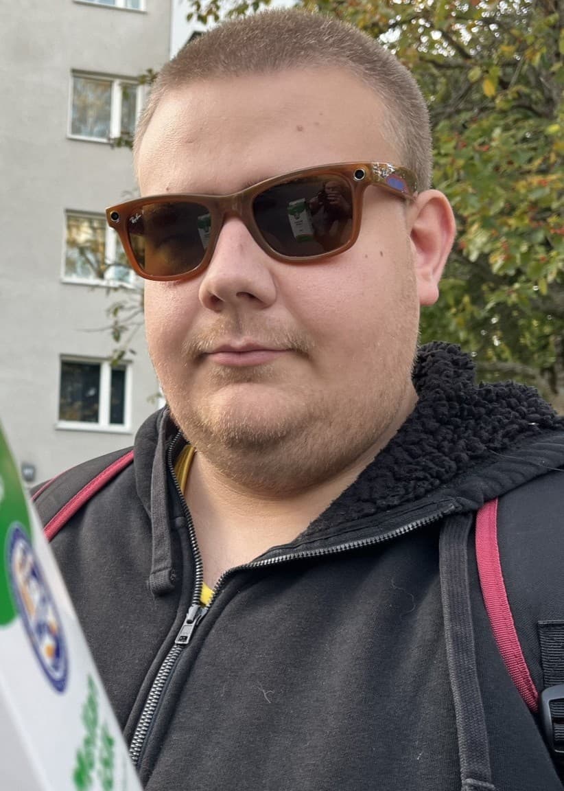 En mjölkförpackning med grön och vit design hålls upp av en man med solglasögon framför en bakgrund med en grå byggnad och träd. 