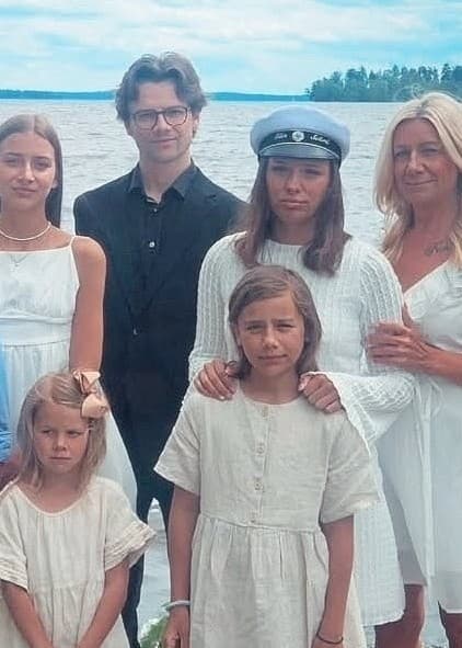 Susanne Zeeck står vid en sjö tillsammans med flera barn och två unga män i studentmössor. Gruppen är klädd i festliga sommarkläder, omgivna av vatten och grönska.