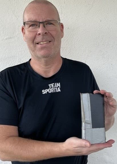 En person klädd i svart T-shirt håller upp en grå rektangulär enhet framför sig, stående utomhus vid en vit husvägg med fönster i bakgrunden.
