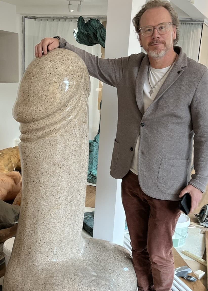 Marcus Skinnar står bredvid en stor granitskulptur i form av en penis, som han köpte i Kina för över 20 år sedan. Rummet är fyllt med andra konstverk och skulpturer.