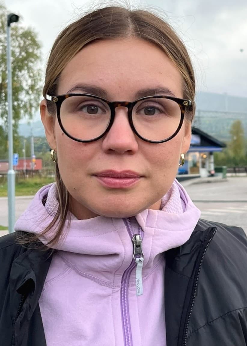 Emma Kuoljok står utomhus på en plats med grönska och moderna byggnader i bakgrunden. Hon bär en lila tröja och svart väst. Molnigt väder med berg syns i fjärran.