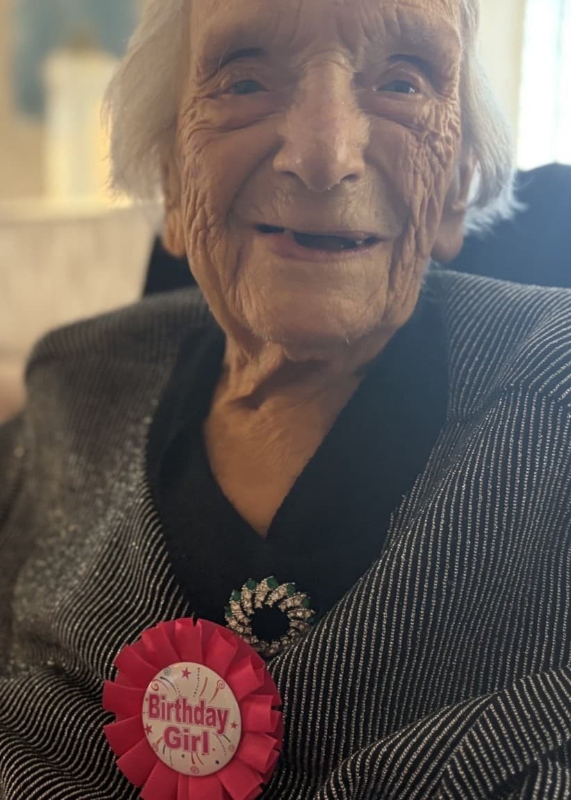 En äldre kvinna sitter i en soffa och bär en grå kofta med en röd rosett som har texten 'birthday girl'. Bakgrunden visar en ljus vardagsrumsmiljö med möbler och tavlor.