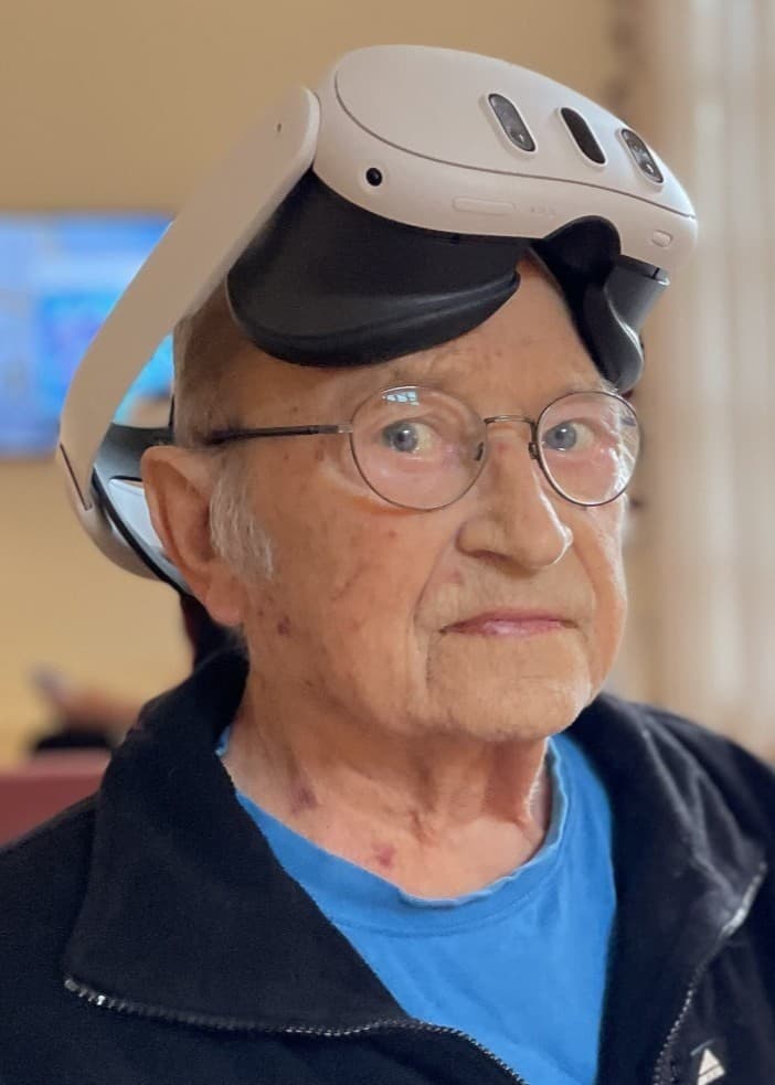 En person med VR-headset på huvudet sitter inomhus i en ljus miljö med stora fönster och växter i bakgrunden. En annan person står bredvid och verkar assistera.