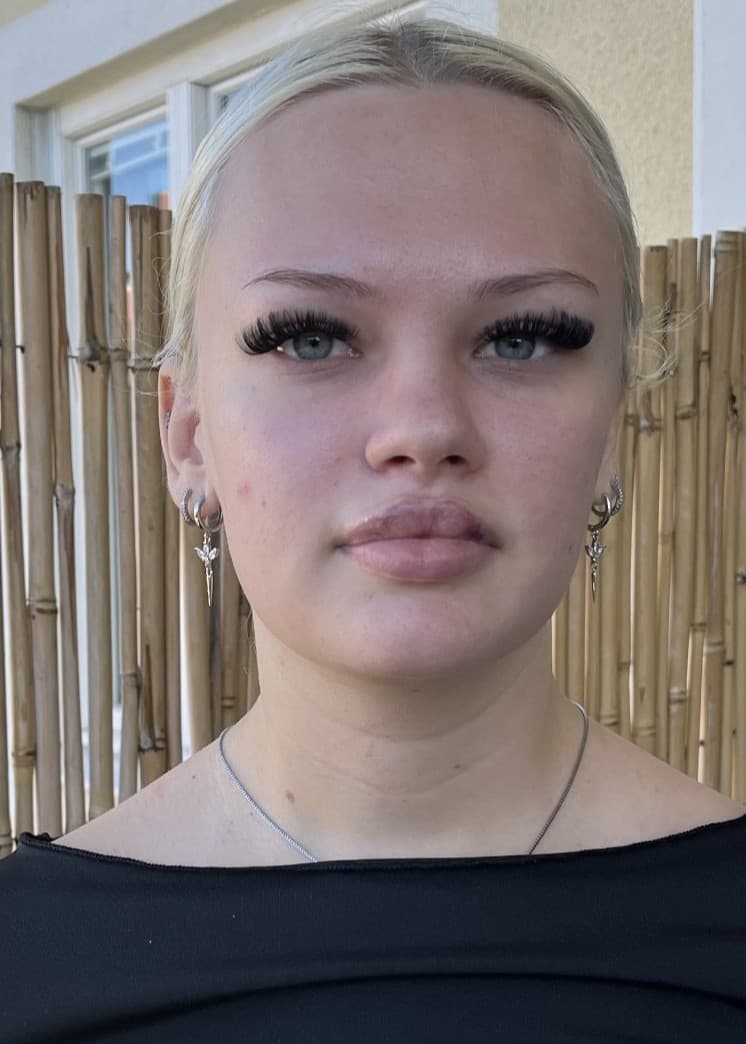 Freya, 18, står framför en bambuskärm utomhus, klädd i en svart topp. Bakgrunden visar en ljus byggnad med fönster.