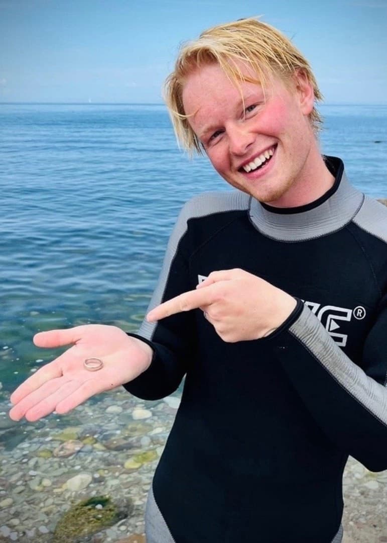 Elias Wärlinge står vid en strand på Öland i våtdräkt och pekar på ett litet föremål i sin hand, som tros vara hittat med en metalldetektor. En närbild av detektorn visas bredvid.