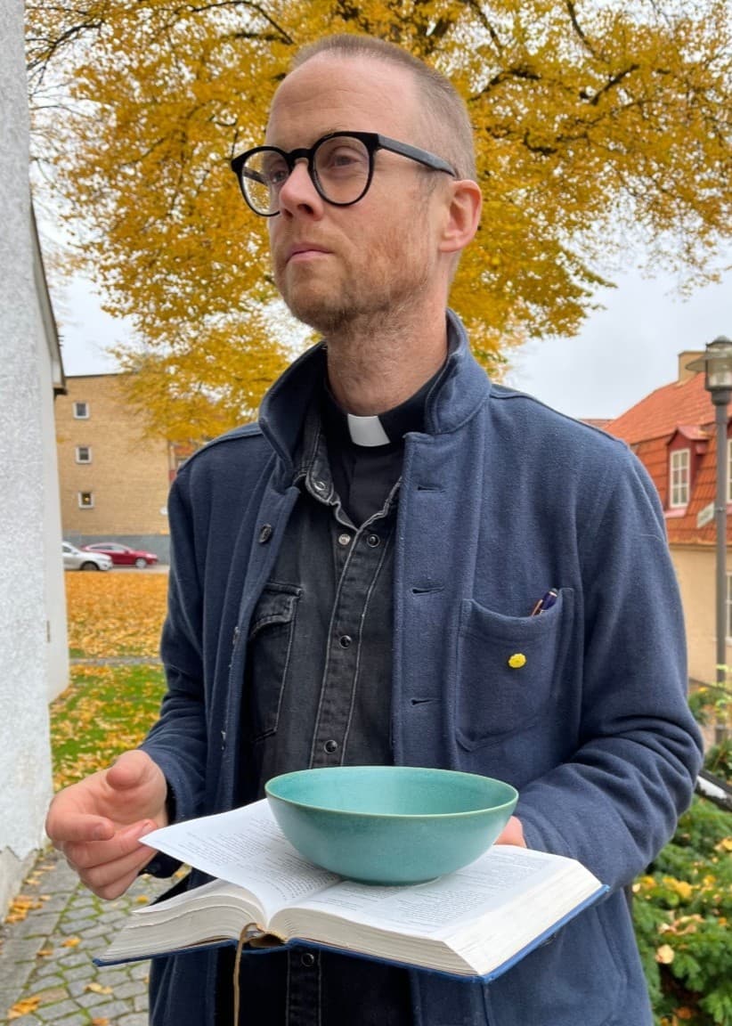 Martin Landgren, präst i Ulricehamn, välsignar en bostad genom att applicera olja på en dörr. Han håller en skål med olja och står utomhus med höstfärgade träd i bakgrunden.