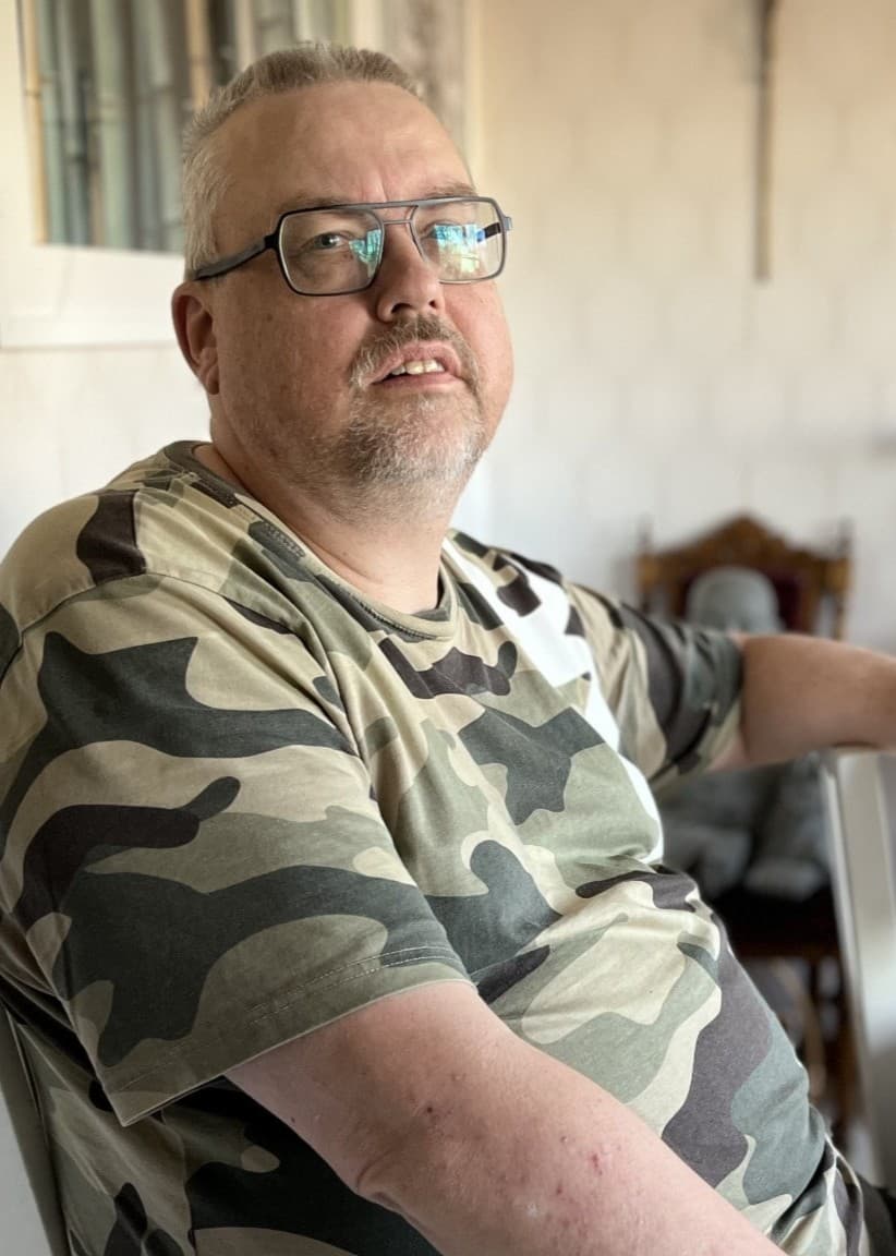 Per Lundberg sitter vid ett träbord i ett rum med religiösa dekorationer, inklusive kors och en staty. Han bär en kamouflagefärgad tröja och vilar sin arm på en stol.