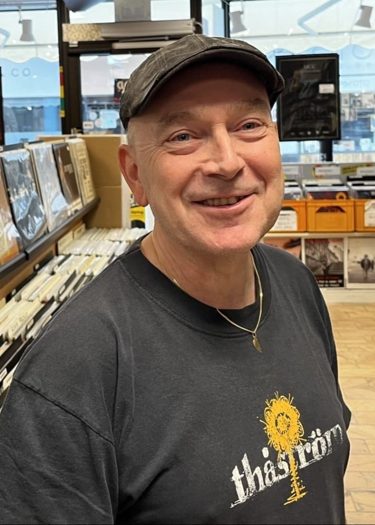 Edvard 'Evve' Nissen står i en skivbutik omgiven av vinylskivor och hyllor fyllda med musik. Han är klädd i en mörk T-shirt med gul text och motiv. Butiken har stora fönster i bakgrunden.
