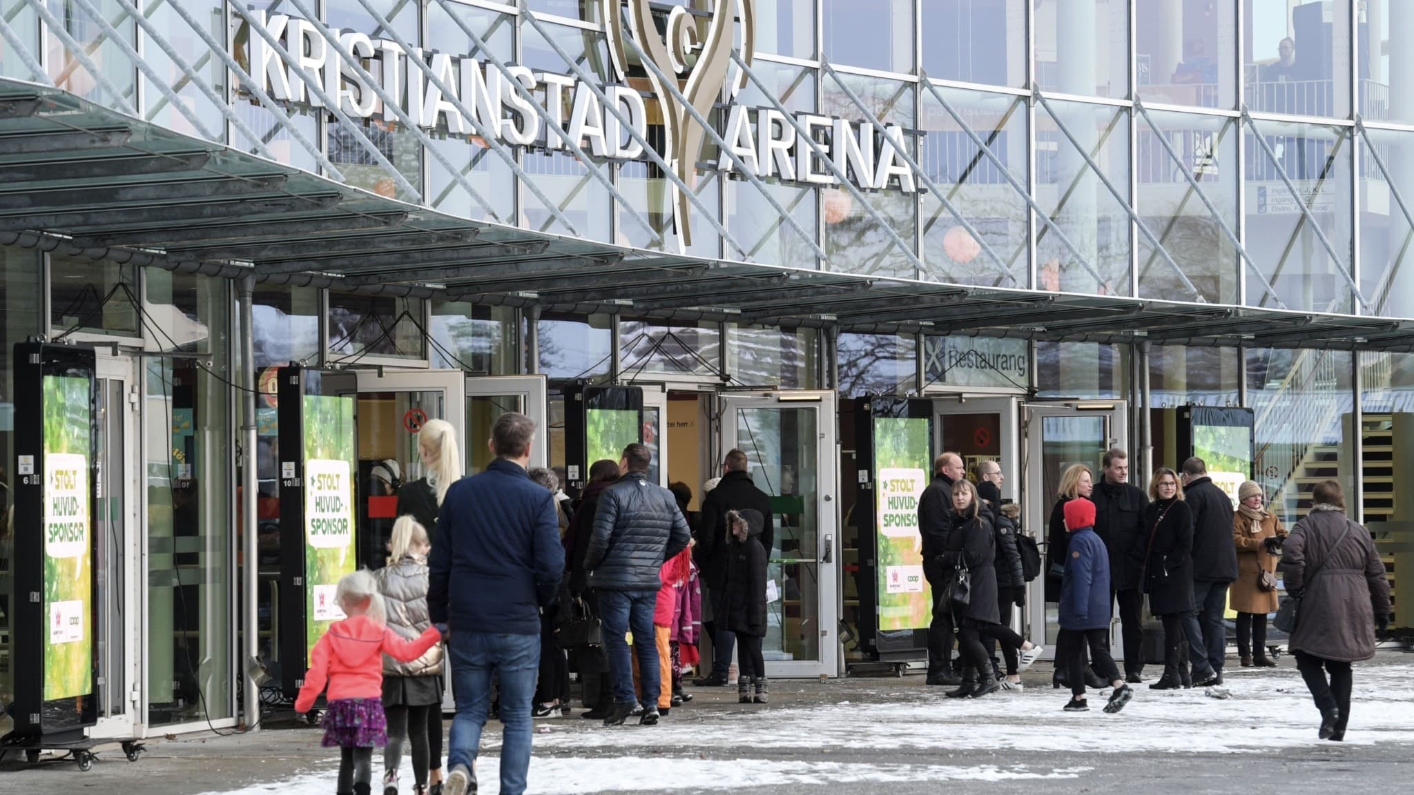 Publik köar vid entrén till Norrköping Arena inför Melodifestivalen. Glasfasaden reflekterar ljus, och flera personer syns med vinterkläder. Stämningen är livlig och förväntansfull.