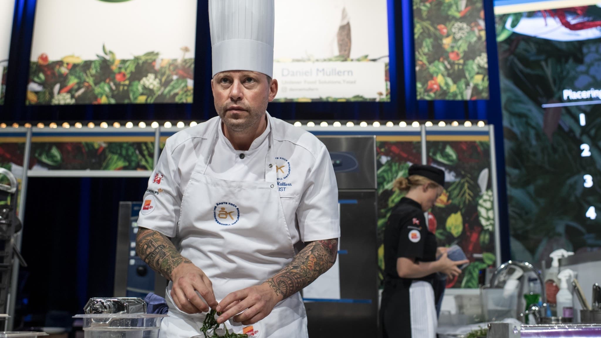 Daniel Müllern, kock från Ystad, står vid en arbetsstation och förbereder mat under en kocktävling. Han bär vit kockrock och högt kockmössa, omgiven av tävlingsutrustning och dekorerad bakgrund.