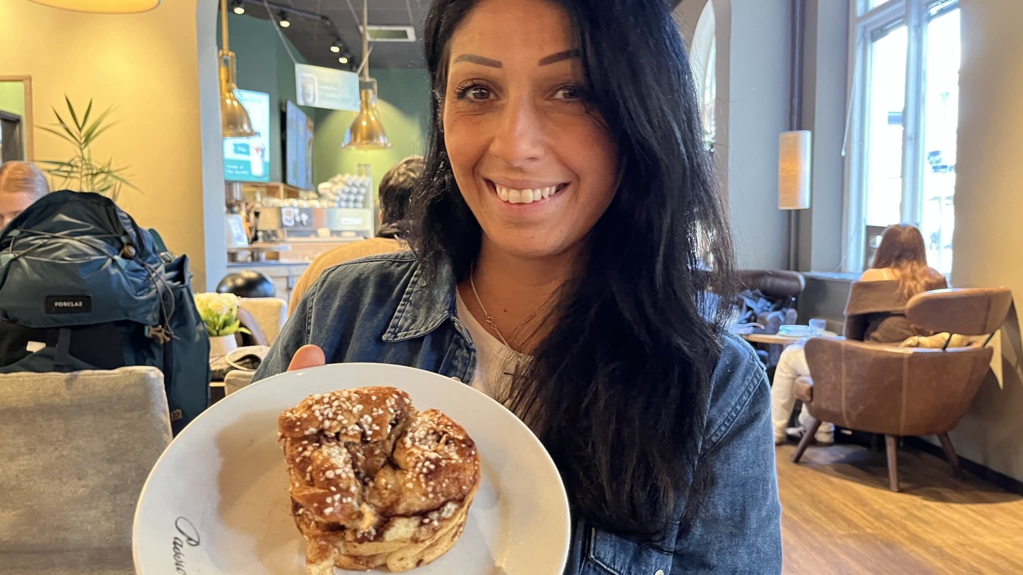 Mayka Gulo, matinfluenser, håller upp en tallrik med en kanelbulle i ett modernt café med stora fönster och dämpad belysning.
