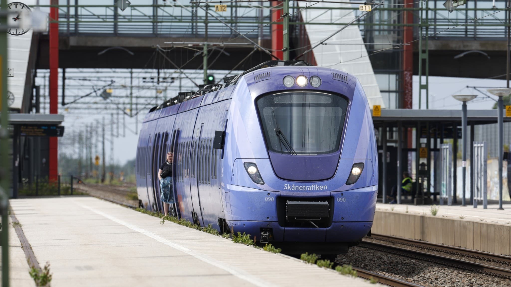 Ett stillastående tåg vid en station Perrongen är tom och en gångbro syns i bakgrunden. 