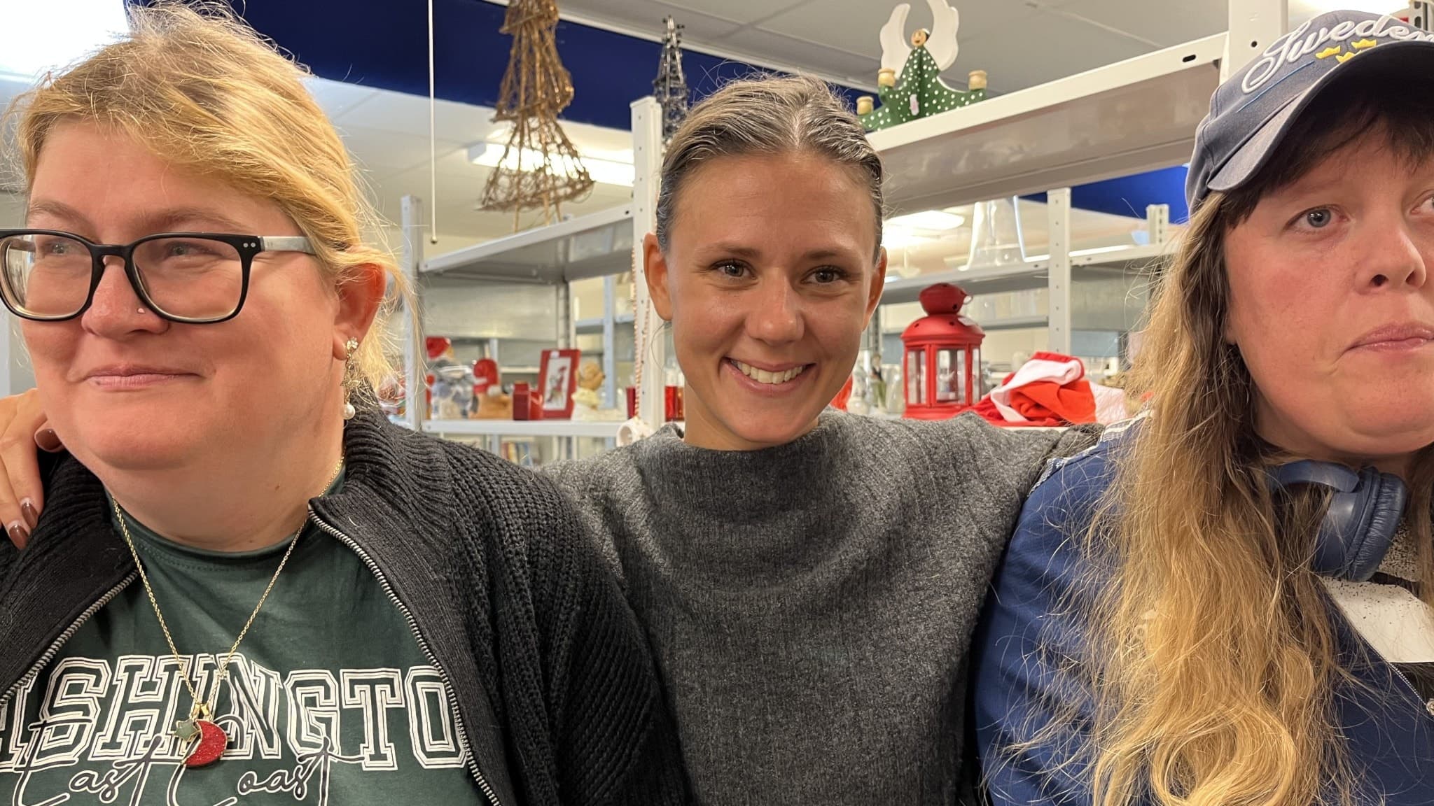 Tre personer står nära varandra i en butiksmiljö med dekorationer i bakgrunden, inklusive röda lyktor och hängande stjärnor. 
