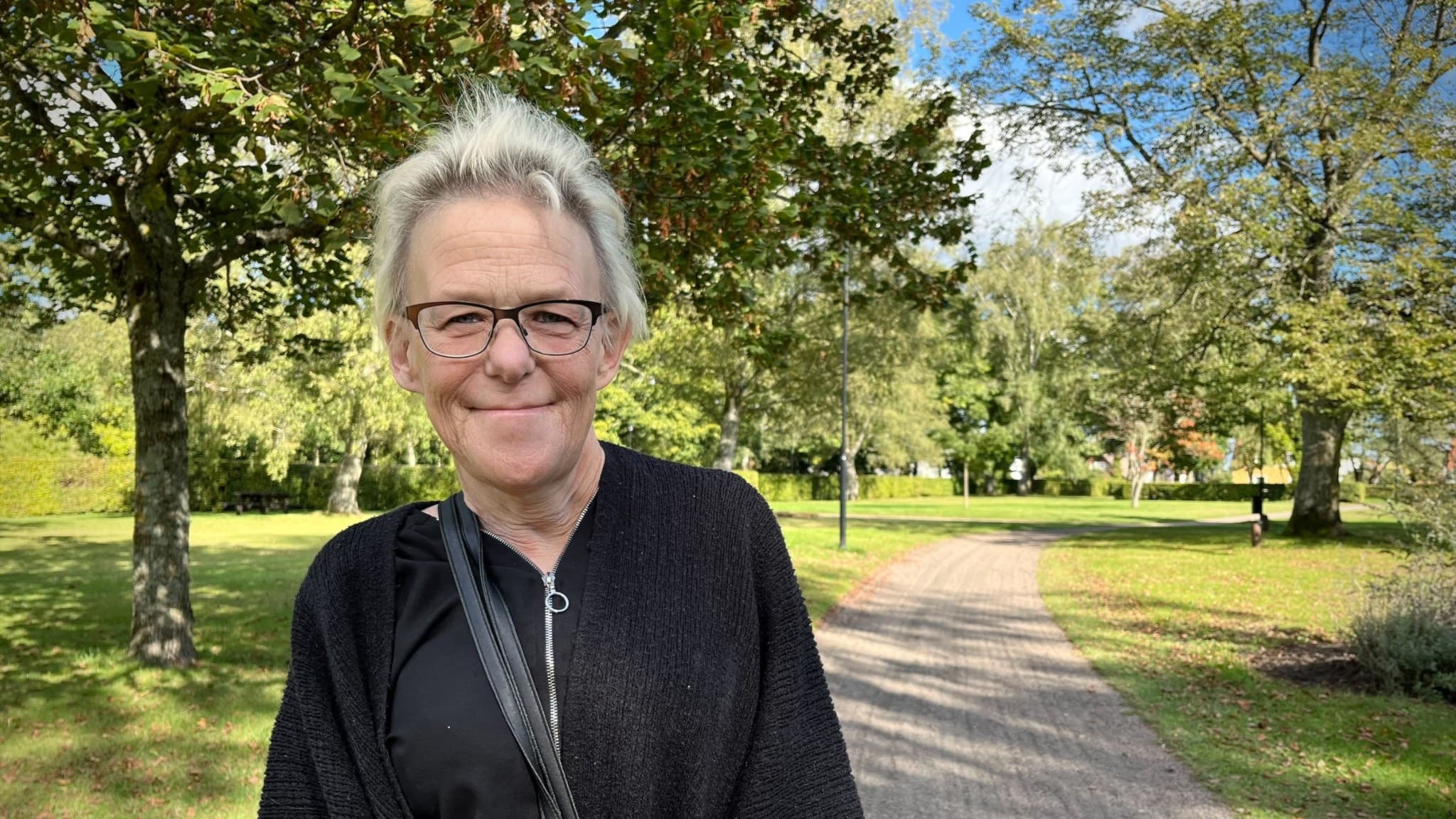 Annika Tydesten från Sverigedemokraterna står i en grönskande park med träd, gräsmattor och en grusväg under en klarblå himmel.