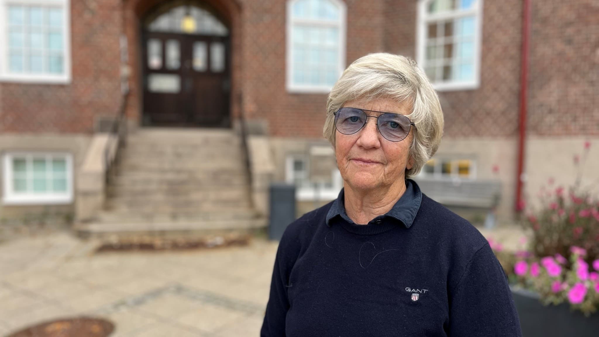 En kvinna står framför en äldre byggnad med tegelvägg och breda trappsteg. På ena sidan syns en blomlåda med färgglada blommor. Kvinnan är klädd i en mörkblå tröja.