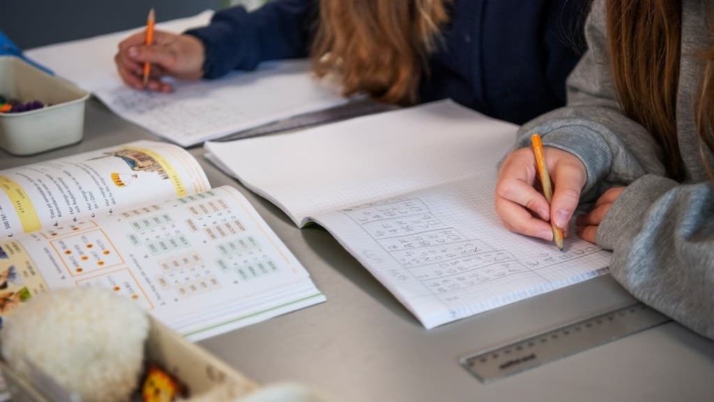 Två barn sitter vid ett bord och skriver i skrivböcker. Framför dem ligger en öppen bok med matematikövningar och en linjal. Bilden illustrerar studier och utbildning.