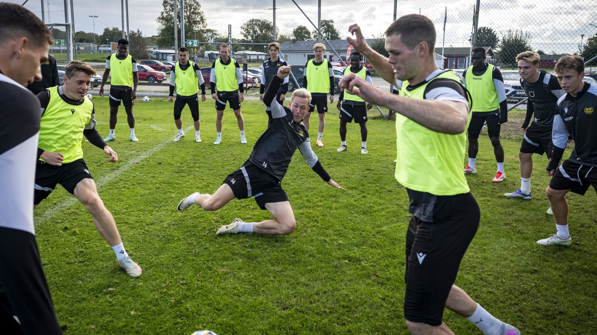 Mjällby AIF-spelarna Elliot Stroud, Herman Johansson och Jacob Bergström tränar intensivt på Strandvallen i Hällevik. Fokuserade spelare jagar bollen på gräsplan under lagövning.