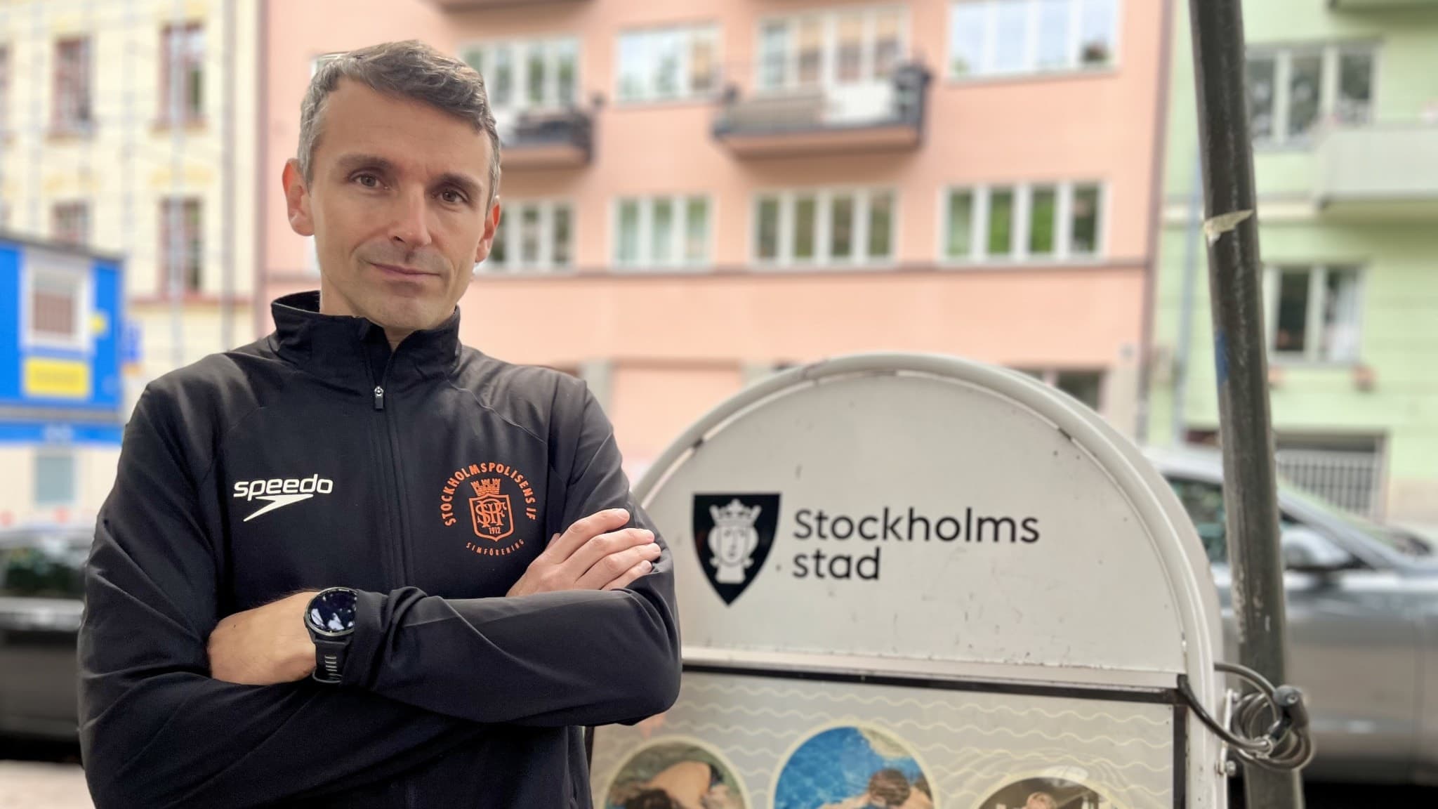 Grega Zora, verksamhetschef för Stockholmspolisens IF Simförening, står utomhus med korsade armar framför en skylt med texten Stockholms stad. Bakgrunden visar bostadshus i en stadsmiljö.