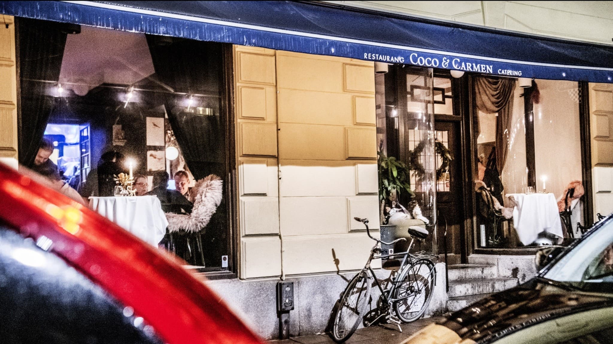 Restaurang Coco & Carmen på Banérgatan i Stockholm med upplysta fönster där gäster syns sitta vid dukade bord. Framför byggnaden står en cykel och bilar parkerade längs gatan.
