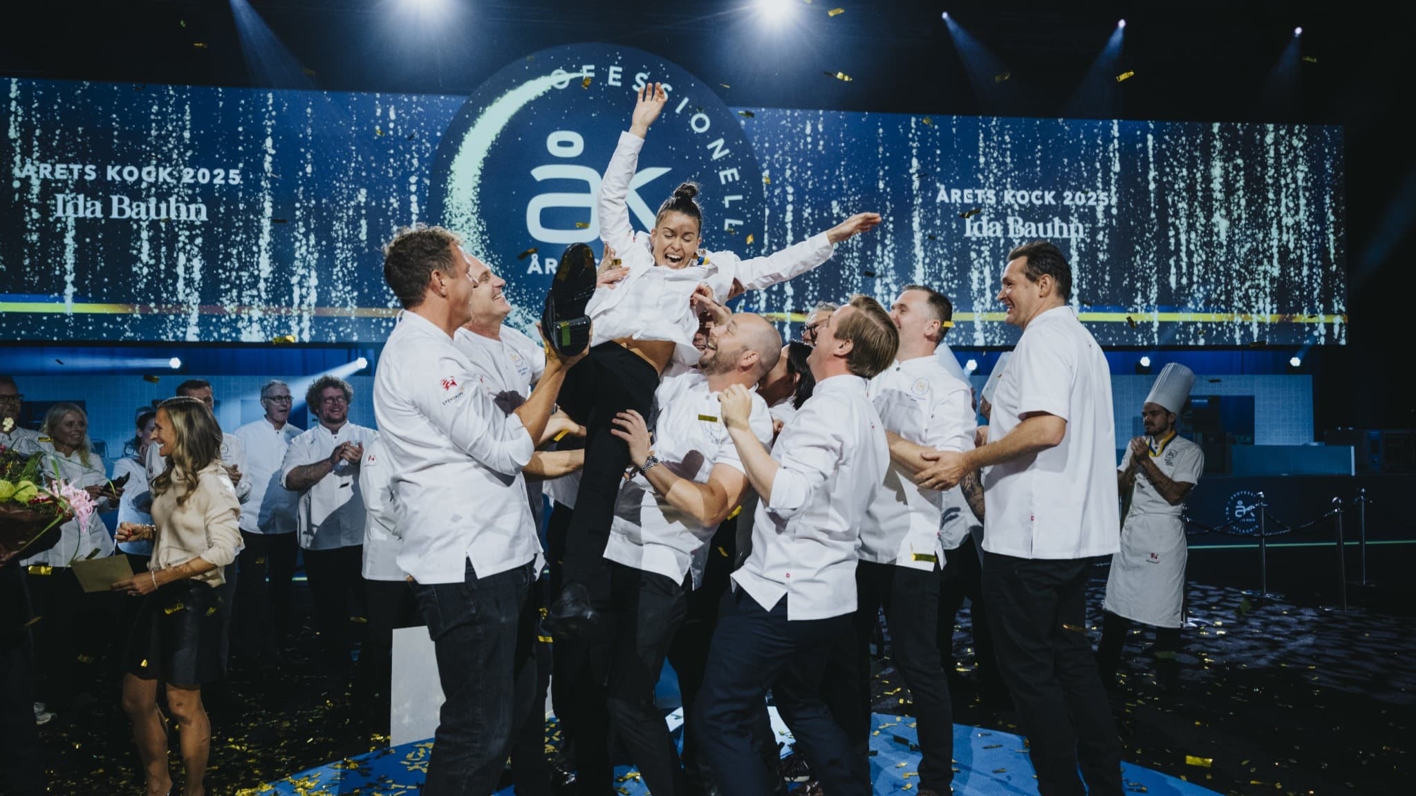 Ida Bauhn firar segern som Årets Kock tillsammans med ett glatt team på scen, omgiven av konfetti och en upplyst bakgrund med texten Årets Kock.