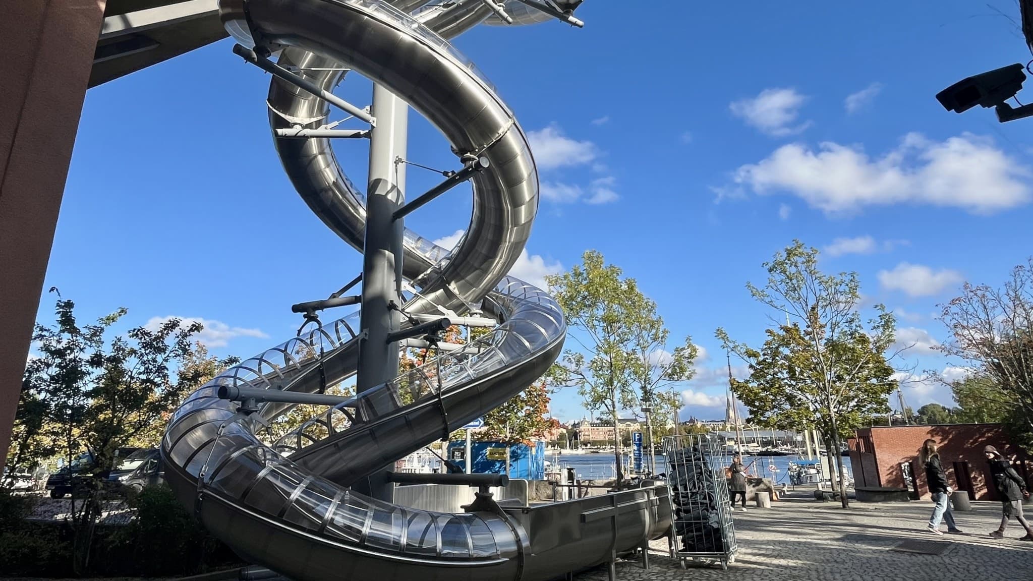 Två 39 meter långa, spiralformade rutschbanor i metall vid Moderna Museet. Soligt väder med blå himmel, omgivna av träd.