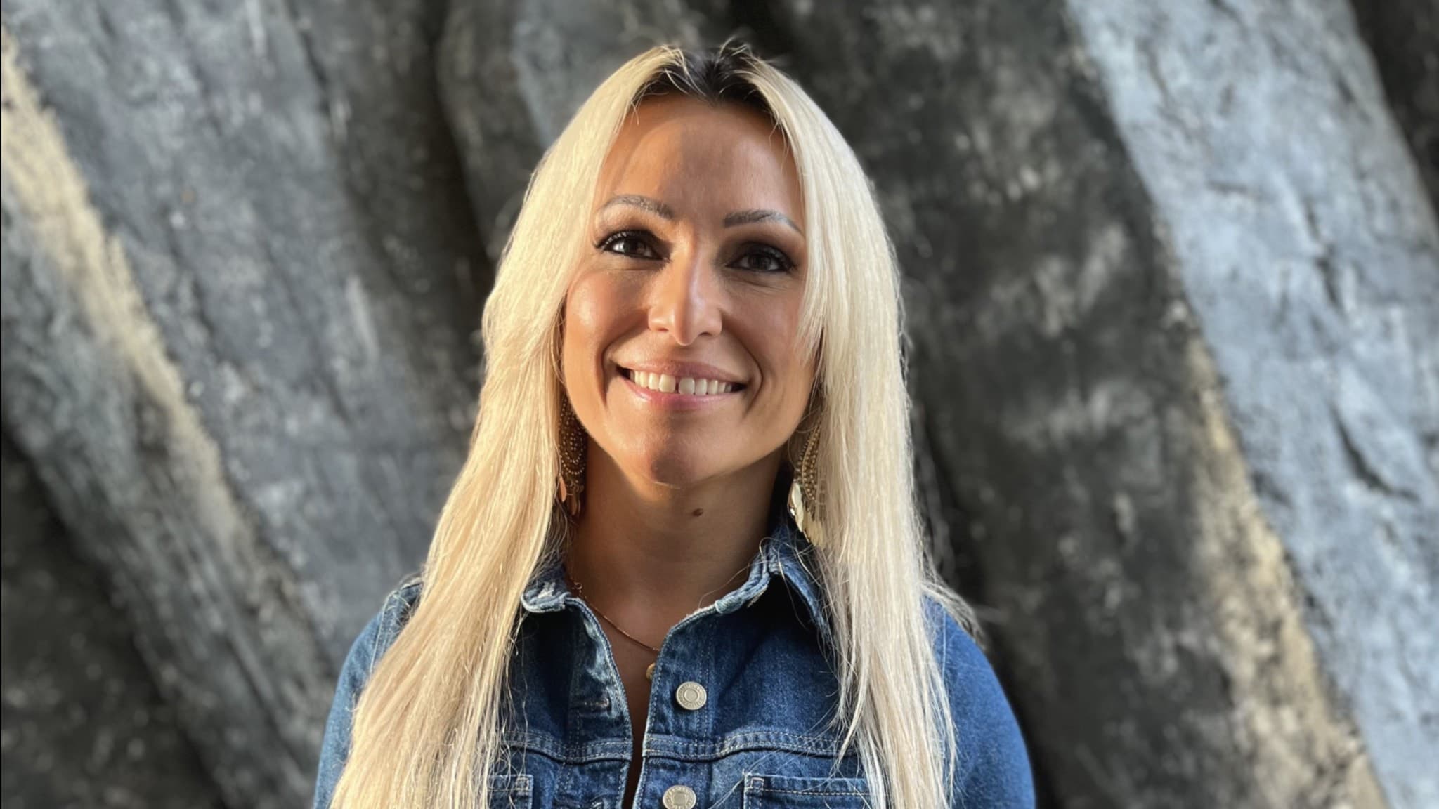 Yury med långt blont hår bär en jeansklänning och står framför en bakgrund. 