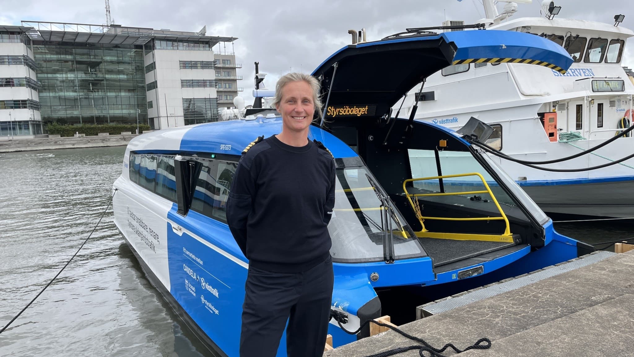 Befälhavare Linda Öhrbom står vid kajen framför den blå och vita flygande färjan i Göteborgs hamn. Färjan testas trots utmanande vindförhållanden som kan påverka framtida rutter.