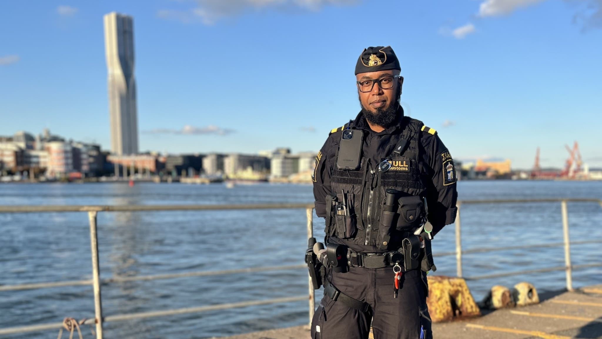 En polis står vid kajen i Göteborg med utsikt över vattnet och den ikoniska skyskrapan Karlatornet i bakgrunden. Solen skiner och byggnader samt kranar syns på andra sidan vattnet.