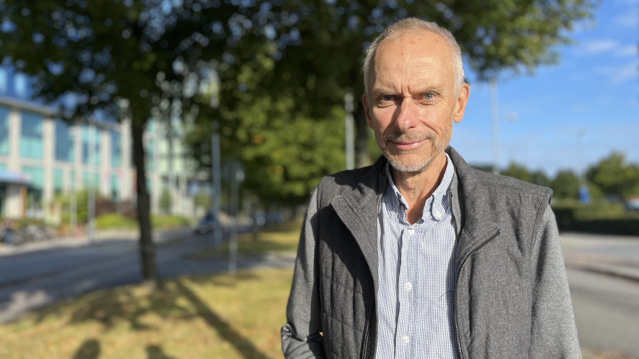 Halvbild på en man i skjorta och grå väst som kisar mot solen. I bakgrunden syns träden mörkgrå vägg.