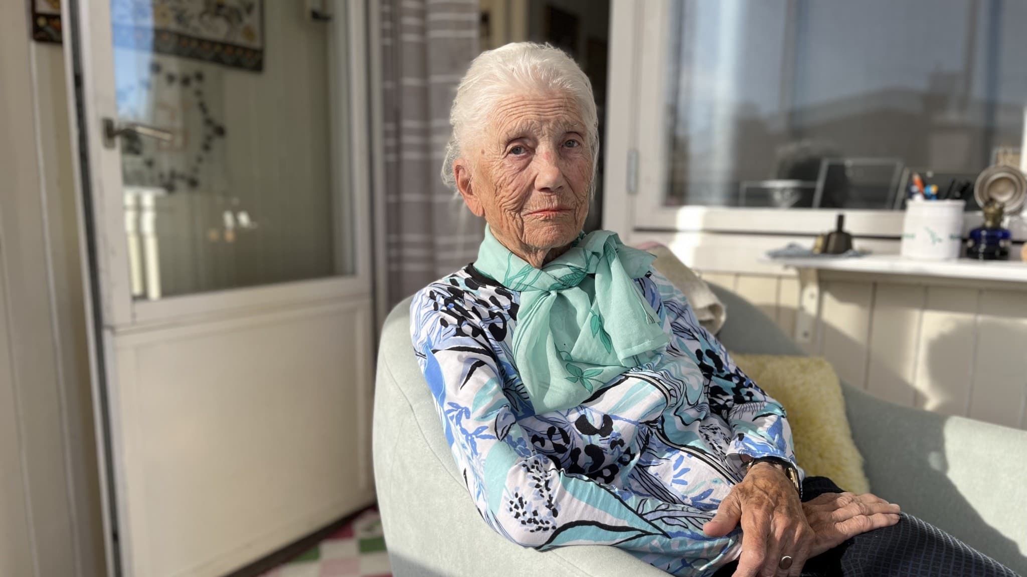 En äldre person sitter i en fåtölj på en solig balkong. Personen bär en mönstrad blus och en grön halsduk. Balkongen är inredd med en dörr, fönster och en färgglad matta.