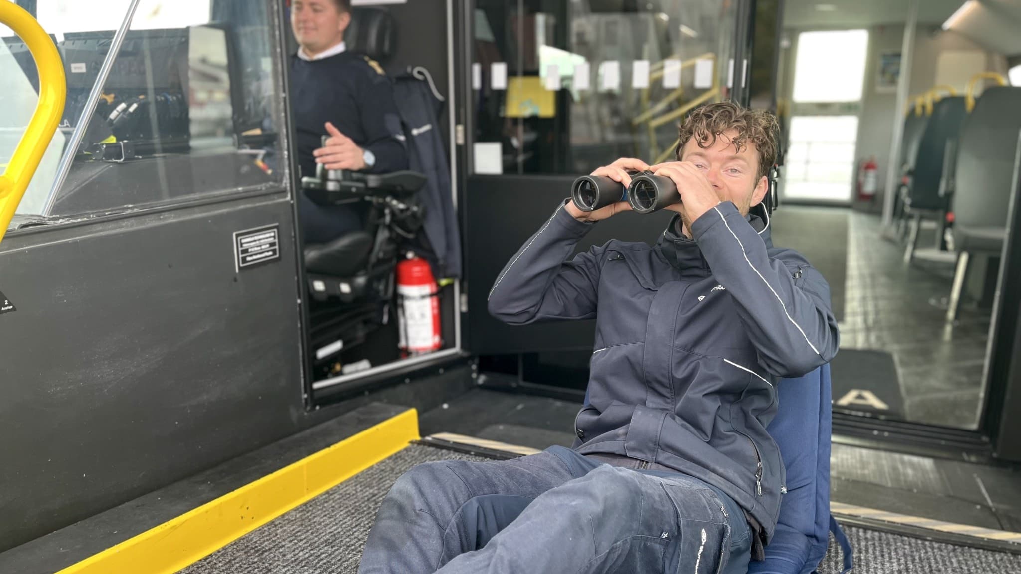 Däcksman Mårten Lundgren sitter i en båt och spanar med kikare efter hummertinor. I bakgrunden syns en annan person vid styrplatsen inne i båtens kabin.