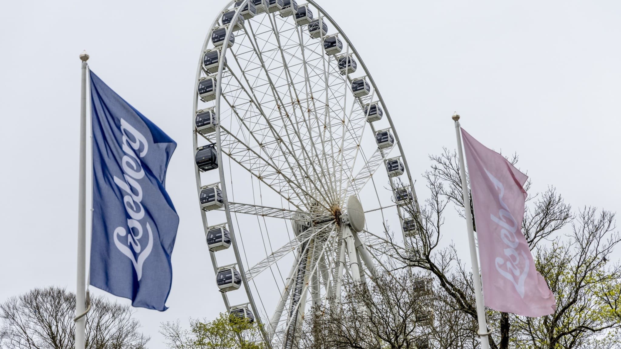 Lisebergshjulet i Göteborg reser sig mot en grå himmel, omgiven av träd och två flaggor med Lisebergs logotyp i blått och rosa, symboliserande nöjesparkens ikoniska attraktioner.